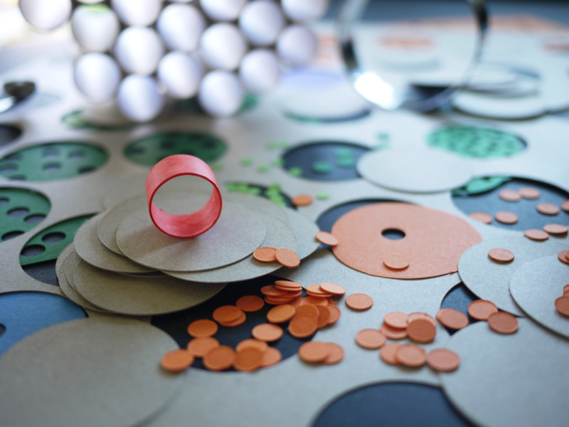 Auf einem Tisch liegen viele runde Formen aus Karton und kleine orangefarbene Kreise. In der Mitte steht ein roter Ring aus Papier. Im Hintergrund liegen weitere Bastelmaterialien.