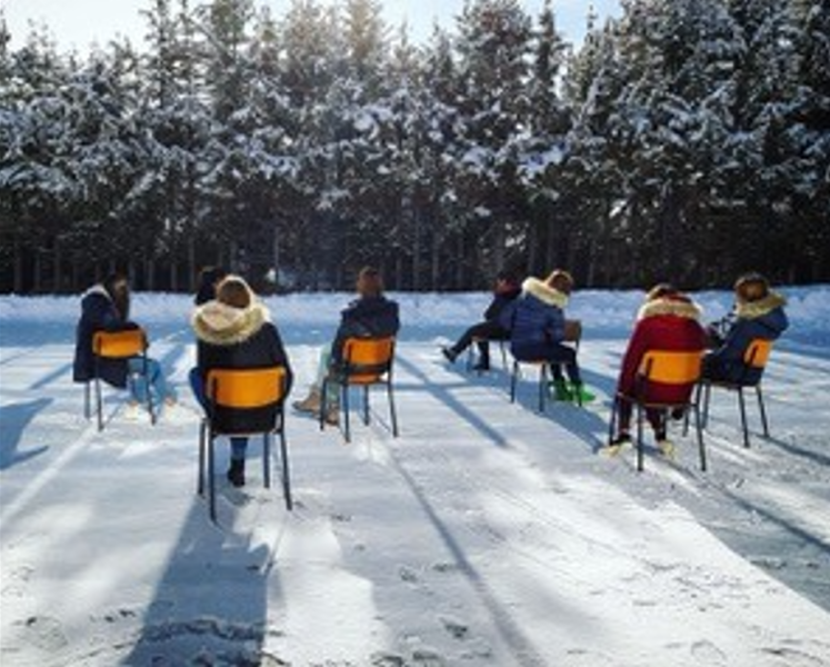 Dieses Bild zeigt eine Gruppe von Menschen, die auf Stühlen in einer Reihe auf einem verschneiten Feld sitzen. Sie blicken auf eine Baumreihe, und die Sonne scheint und wirft lange Schatten hinter sie. Die Umgebung lässt auf eine winterliche Umgebung schl