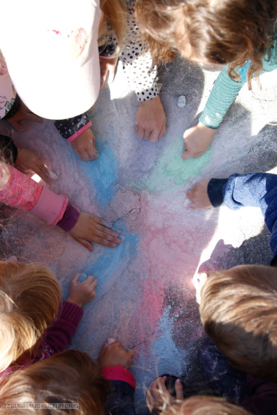 Das BIld zeigt Kinderhände im Kreis, die mit bunten Kreiden Kreiden auf den Boden malen.