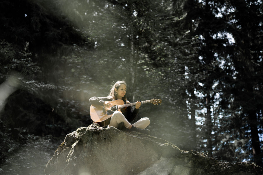 Emelyne sitzt mit ihrer Gitarre im Wald auf einem grossen Stein.