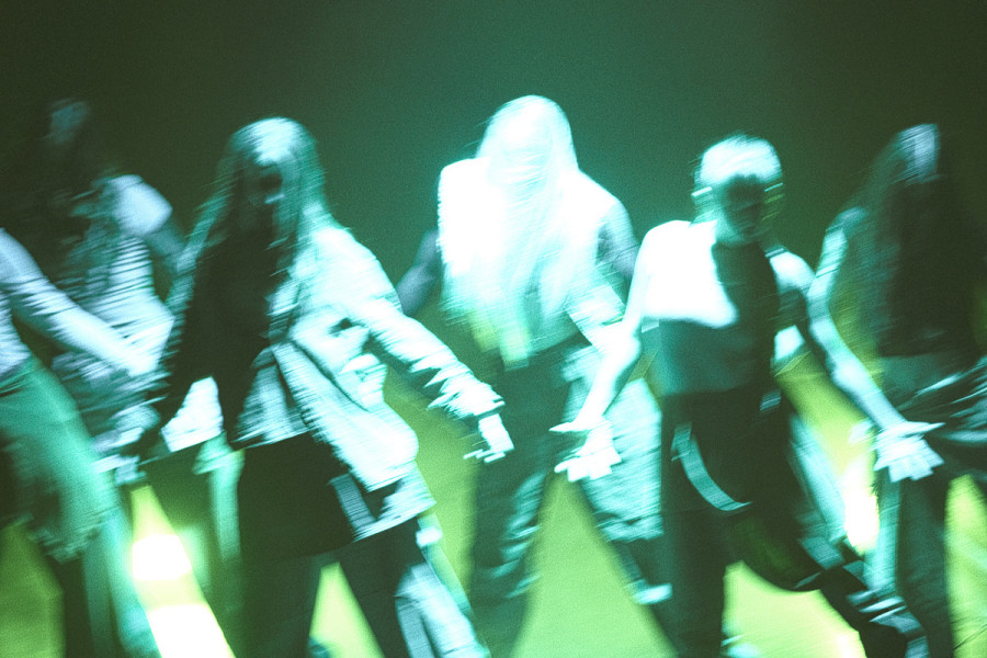 Auf dem Bild sind mehrere Menschen auf einer Bühne zu sehen. Sie bewegen sich gleichzeitig, vermutlich in einer Tanzperformance.  Die Figuren sind durch Bewegung unscharf, sodass ihre Körper leicht verschwimmen. Das Licht ist grünlich und kommt von unten 