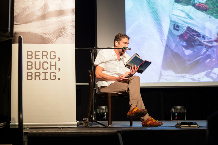 Ein Mann sitzt vor dem Plakat von BergBuchBrig auf der Bühne und liest aus einem Buch vor.