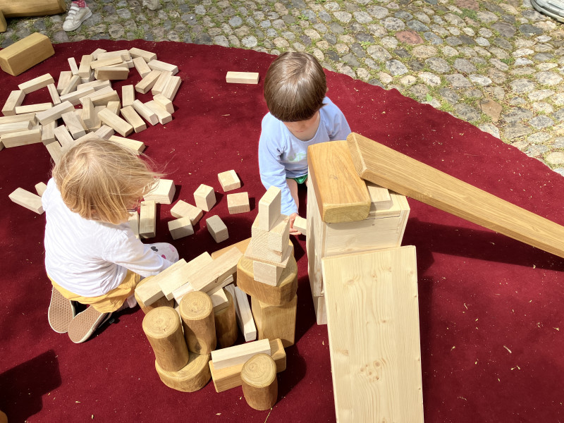 Zwei Kinder spielen draussen auf einem roten Teppich. Sie bauen gemeinsam mit grossen Holzklötzen und Holzbrettern. Um sie herum liegen viele weitere Holzklötze.