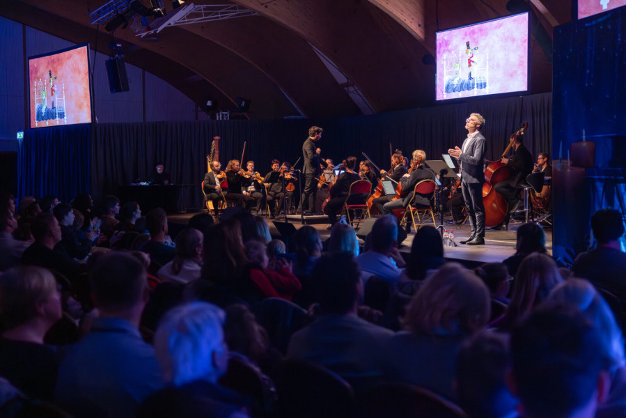 Das Bild zeigt ein Familienkonzert in einem großen Saal. Viele Menschen sitzen im Publikum und schauen nach vorne auf die Bühne.  Auf der Bühne spielt ein kleines Orchester. Man sieht Musikerinnen und Musiker mit Geigen, einem Kontrabass und einer Harfe. 