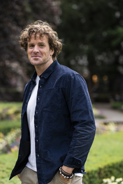Ein Mann steht draußen in einem Garten oder Park. Er schaut leicht zur Seite und lächelt freundlich. Der Mann hat lockige, braune Haare.  Er trägt ein dunkelblaues Hemd über einem weißen T-Shirt. Dazu trägt er eine helle Hose. An seinem Handgelenk sind me