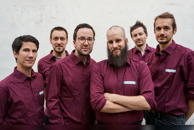 Die Band trägt ein rotes Hemd. Es sind 6 Männer auf dem Bild zu sehen. 
