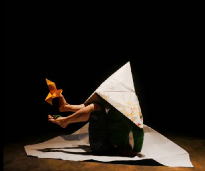 Das Bild zeigt eine Person, die mit einer großen Papierstruktur bedeckt ist, die möglicherweise einem Zelt oder einem Boot ähnelt. Die Person hält ein gelbes Origami-Stück mit den Füßen, wobei ein weiteres kleines Origami-Stück in der Nähe ihrer Zehen sic