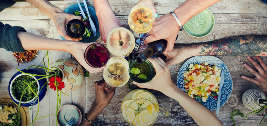 Sechs Arme von verschiedenen Personen aus aller Welt stossen mit bunten Getränken gemeinsam an. Der Tisch darunter ist mit verschiedenen Speisen gedeckt.