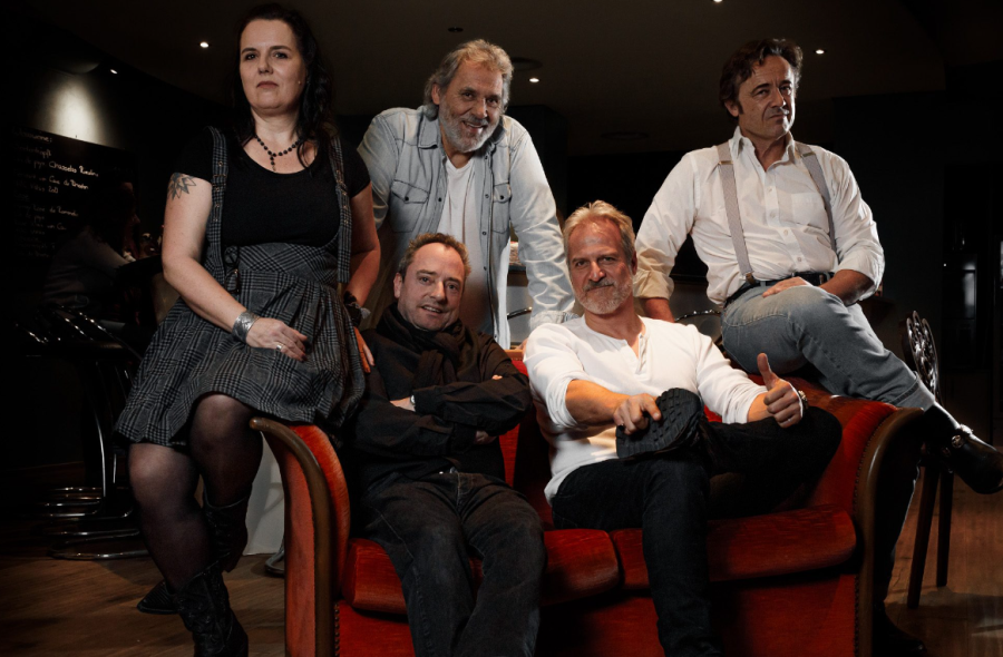 Die Band sitzt auf eine Sofa. Vier Musiker und eine Musikerin in weiss und schwarz gekleidet