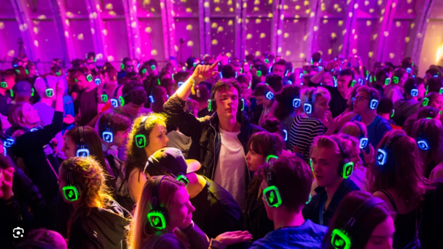 Auf dem Bild ist eine grosse Menschenmenge zu sehen, die zusammen tanzt. Die Szene findet in einer Halle mit buntem Licht statt. Das Licht ist vor allem pink und violett, mit vielen kleinen gelben Lichtpunkten an der Decke.  Alle Menschen tragen grosse Ko