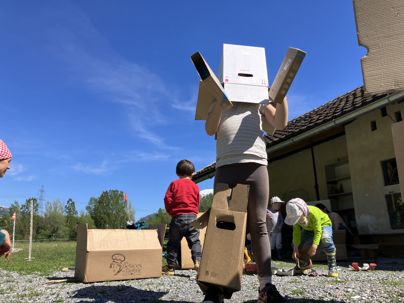 Kinder spielen draussen mit grossen Kartonschachteln. Ein Kind trägt eine Schachtel auf dem Kopf und hält zwei Kartonstücke in die Höhe. Im Hintergrund spielen weitere Kinder vor einem Haus. Die Sonne scheint.
