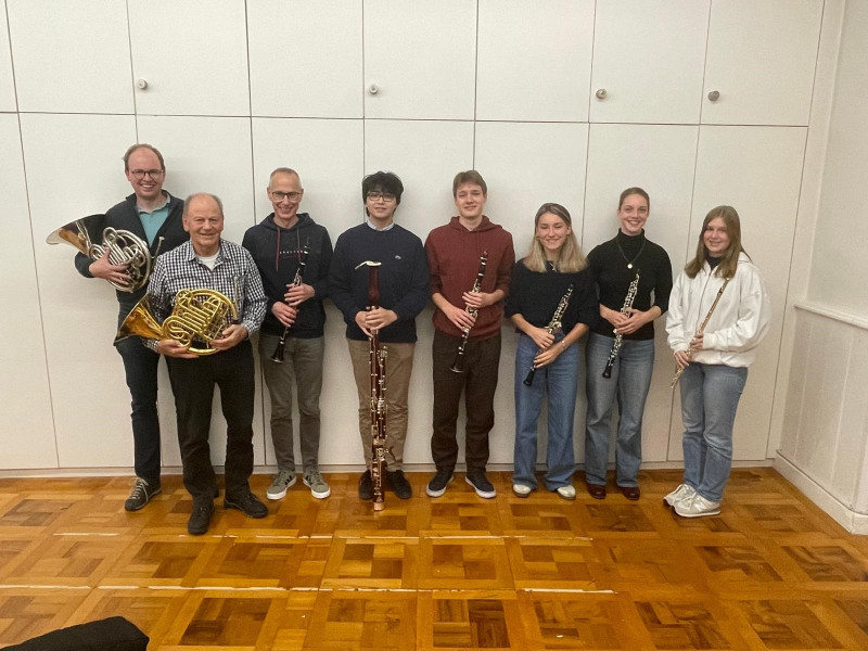 acht Musikerinnen und Musiker der Serenata Oberwallis stehend an einer weissen Wand mit ihren Instrumenten.