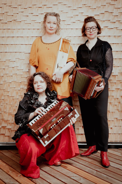Auf dem Bild sind drei Frauen mit Musikinstrumenten zu sehen. Sie posieren vor einem strukturierten Holzhintergrund. 