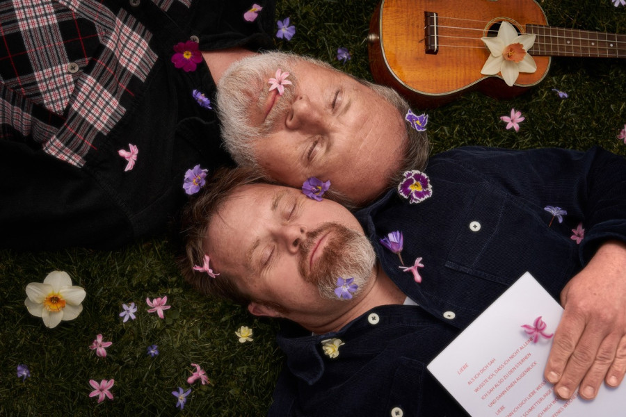 Dieses Bild zeigt zwei Männer, die im Gras liegen, umgeben von verstreuten Blumen. Sie wirken entspannt und haben Blumen auf sich, wobei einer von ihnen ein Blatt Papier hält. In der Nähe steht auch eine Ukulele, die zu einer ruhigen und künstlerischen Ko
