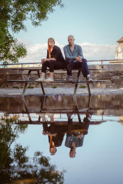 Der Sänger Manuel Pollinger und die Piantistin Meta Červ sitzen an einem See auf einer Bank. Ihr Spiegelbild findet sich im See wieder.
