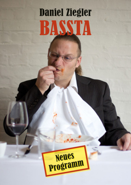 Daniel Ziegler mit Ross-Schwanz und Anzug sitzt an einem Tisch. Er hat eine dreckige Serviette umgebunden und einen leeren Teller mit Tomatensaucen-Resten vor sich .