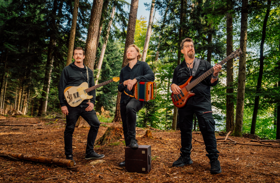 Dieses Bild zeigt drei Musiker in einem Wald. Alle tragen schwarze Kleidung. Links hält jemand eine E-Gitarre, in der Mitte steht jemand mit einem Akkordeon auf einer Box, und rechts spielt jemand einen Bass.